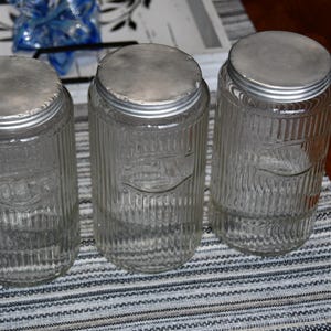 May include: Three clear glass jars with ribbed sides and metal lids. The jars have a vintage appearance and are arranged on a striped surface. The jars are empty and ready to be filled.