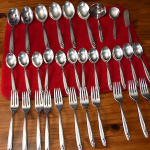 May include: A collection of stainless steel silverware, including spoons, forks, and serving utensils, arranged on a red cloth. The silverware has a simple, elegant design, reflecting a classic aesthetic. The arrangement suggests a formal dining setting.