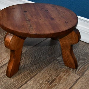 May include: A small, round wooden stool with a dark brown stained finish. The stool features a smooth, circular top surface with visible wood grain patterns and is supported by three curved, sturdy legs that extend outward from the base.