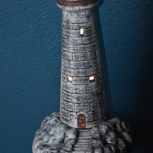 May include: A decorative lighthouse figurine with a grey stone-like exterior. The lighthouse has a brown top and base, with small windows that appear to be illuminated. The base is a rocky formation. The figurine is approximately 25 cm tall.