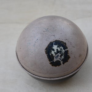 May include: A round, silver-colored container with a black base. The container has a worn, faded sticker with a white design on it.