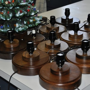 May include: A collection of wooden lamp bases with black lamp sockets. The bases are round and have a dark brown finish. A ceramic Christmas tree with colorful lights is visible in the background.