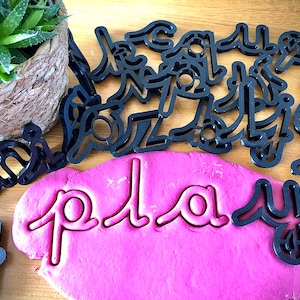 May include: Pink play dough with the word "play" pressed into it using black cookie cutters. The cookie cutters are scattered around the play dough.