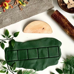 May include: A green play dough mat with six rectangular indentations. The mat is surrounded by green leaves and a wooden log. There are also small plastic frogs on the log.