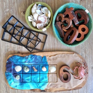 May include: A wooden tray with blue and brown play dough, a wooden number six, a red crab, and a grid of squares filled with seashells.