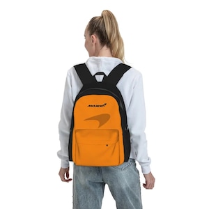 Könnte beinhalten: Ein leuchtend orangefarbener Rucksack mit schwarzen Trägern und Besätzen, der das schwarze McLaren-Logo sowie eine dezente, tonale McLaren-Grafik auf der Vordertasche aufweist. Der Rucksack verfügt über ein Hauptfach und eine Fronttasche.