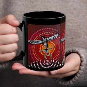 May include: Black ceramic mug featuring an Aboriginal art design. The artwork depicts a central figure with outstretched arms, surrounded by a pattern of red, orange, and yellow dots, with a stylized bird above. The mug is held by a person.
