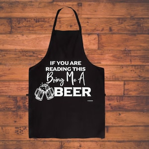 May include: Black apron with white text that reads "IF YOU ARE READING THIS Bring Me A BEER" and a graphic of two beer mugs. The apron is hanging against a wooden background.