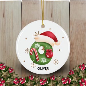 May include: White ceramic Christmas ornament with a green letter O decorated with snowflakes, a candy cane, and a red gift bag. The letter O is wearing a red Santa hat. The ornament has the name "OLIVER" printed below the letter.
