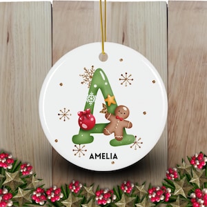 May include: A white ceramic Christmas ornament with a green letter A decorated with gold glitter and snowflakes. A gingerbread man with a red bow tie is on the right side of the letter. The ornament has the name Amelia written below the letter.