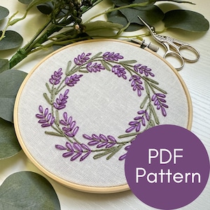 May include: A close-up of a lavender wreath embroidery pattern in a wooden embroidery hoop. The pattern is on a white linen fabric. The text "PDF Pattern" is on a purple circle in the lower right corner of the image.