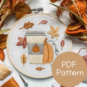 May include: Embroidered hoop art featuring a coffee cup with a pumpkin inside, a pumpkin, and autumn leaves in shades of orange, brown, and green. The hoop is on a white surface with the text "PDF Pattern" in a brown circle.