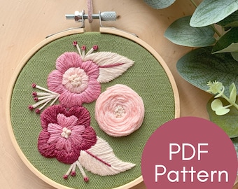 May include: Embroidered floral hoop art featuring pink and burgundy flowers and leaves on a green background. The hoop is hanging from a brown leather cord. The text "PDF Pattern" is on a pink circle in the bottom right corner of the image.
