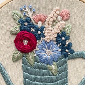 Op de afbeelding: Een blauwe, geborduurde gieter gevuld met een boeket van roze, rode, blauwe en witte bloemen. De bloemen zijn geborduurd in verschillende steken, waaronder satijnsteek, Franse knopen en zaadsteek.