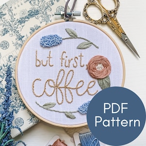May include: Embroidered hoop art with the text "but first, coffee" in brown thread. The hoop is decorated with blue and pink flowers and green leaves.