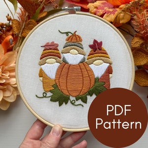 以下が含まれることがあります： 3人のノームが秋の色の帽子をかぶり、中央に大きなカボチャが置かれた刺繍のフープアート。ノームは緑の葉で囲まれています。フープは木でできており、白い布の背景があります。画像の右下隅に「PDF Pattern」というテキストが表示されています。