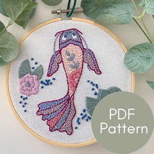 May include: Embroidered hoop art featuring a pink and blue fish with a floral design. The hoop is made of wood and has a white fabric background. The text "PDF Pattern" is visible in the lower right corner of the image.