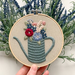 Op de afbeelding: Borduurwerk in een ronde hoepel met een blauwe gieter gevuld met een kleurrijk boeket bloemen. De bloemen zijn roze, rood, blauw en groen.