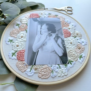 May include: A white embroidery hoop with a floral design in shades of pink, white, and green. A black and white photo of a person is centered in the hoop.