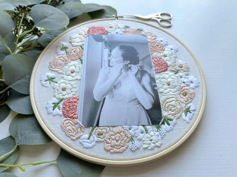 May include: A white embroidery hoop with a floral design in shades of pink, white, and green. A black and white photo of a person is centered in the hoop.