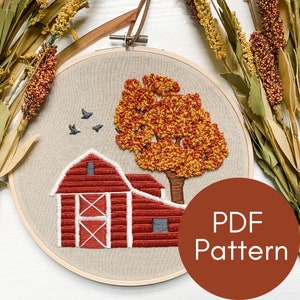 May include: Embroidered hoop art featuring a red barn with a white door and a fall-colored tree with orange and yellow leaves. The hoop is on a white background with dried grasses. The text "PDF Pattern" is on a brown circle.