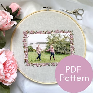Puede incluir: Un aro de bordado de punto de cruz con un borde floral y una foto de una pareja tomándose de la mano en un parque. El aro está sobre una superficie blanca con unas tijeras y una flor rosa. El texto "PDF Pattern" está en un círculo rosa.