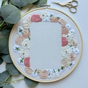 May include: Embroidered hoop art with a floral wreath design. The wreath is made up of pink, white, and cream flowers with green leaves. The hoop is made of wood and has a natural finish.