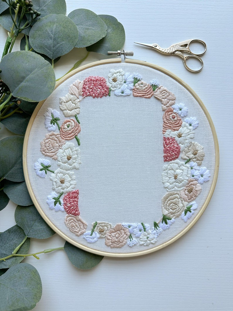 May include: Embroidered hoop art with a floral wreath design. The wreath is made up of pink, white, and cream flowers with green leaves. The hoop is made of wood and has a natural finish.