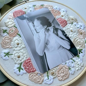 May include: A black and white photo of a person wearing a white dress is framed in a round embroidery hoop. The hoop is decorated with embroidered flowers in shades of pink, white, and beige.