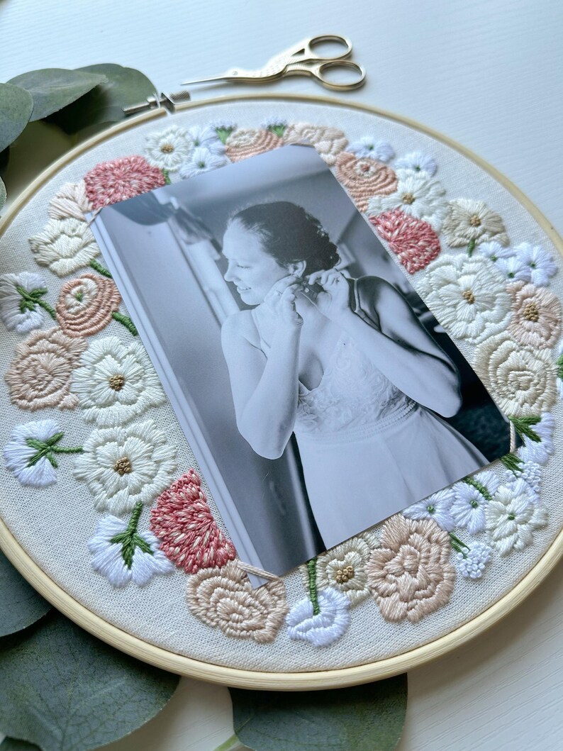 May include: A black and white photo of a person wearing a white dress is framed in a round embroidery hoop. The hoop is decorated with embroidered flowers in shades of pink, white, and beige.
