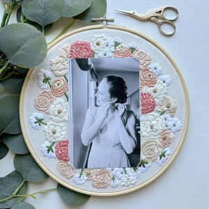 May include: A white embroidery hoop with a floral design in shades of pink, white, and beige. A black and white photo of a person is centered in the hoop.