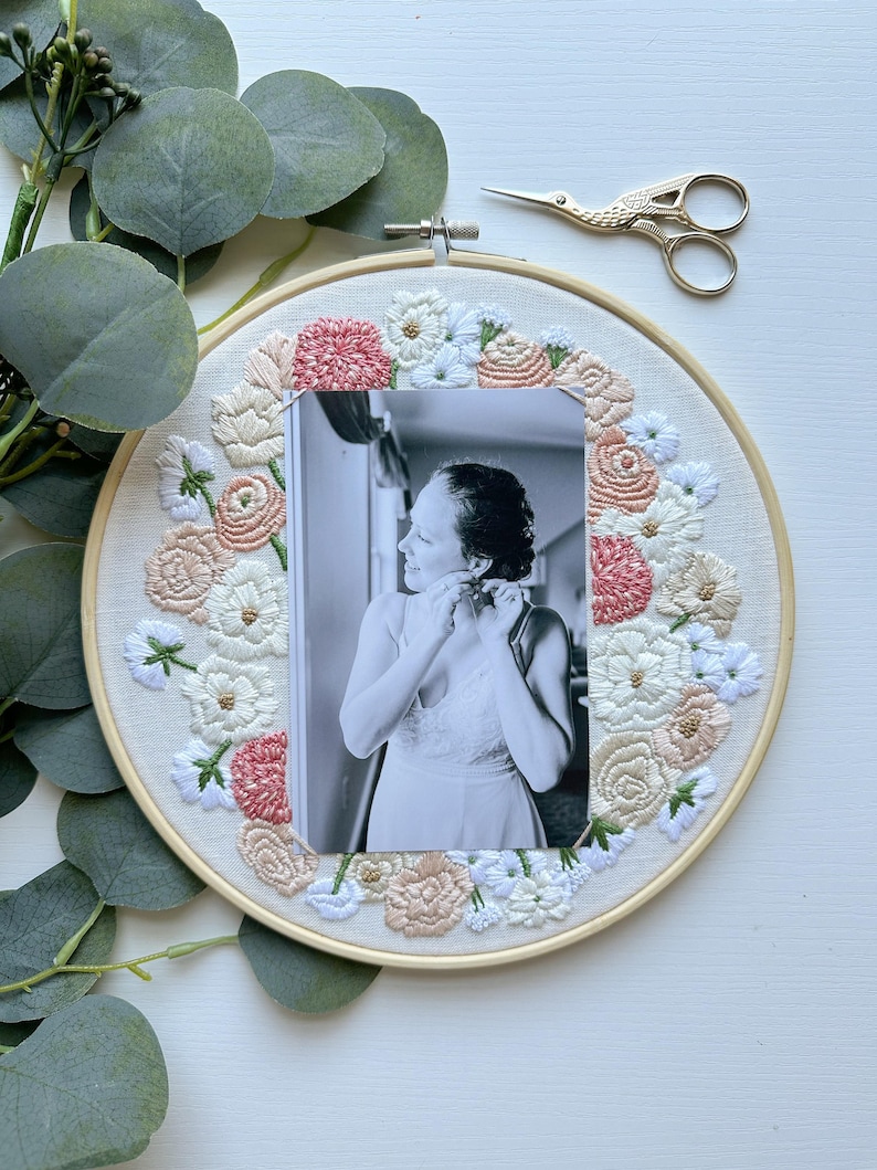 May include: A white embroidery hoop with a floral design in shades of pink, white, and beige. A black and white photo of a person is centered in the hoop.