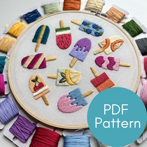 May include: A close-up of a white embroidery hoop with a variety of colorful embroidered popsicles. The hoop is surrounded by spools of embroidery floss in various colors. The text "PDF Pattern" is written in white on a teal circle.