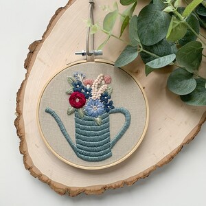 Op de afbeelding: Borduurwerk in een hoepel met een blauwe gieter gevuld met een boeket van roze, blauwe en rode bloemen. De hoepel is van hout en heeft een natuurlijke afwerking.