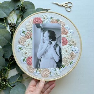 May include: A white embroidery hoop with a floral design in shades of pink and white. A black and white photo of a person is centered in the hoop.
