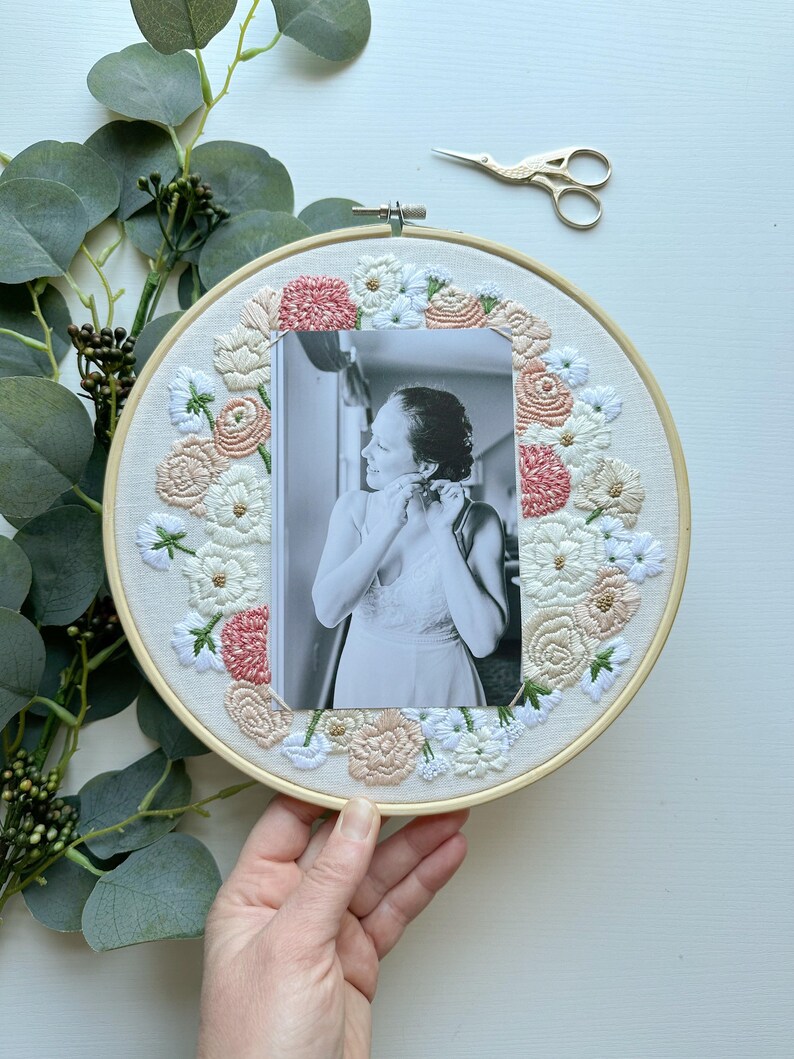 May include: A white embroidery hoop with a floral design in shades of pink and white. A black and white photo of a person is centered in the hoop.