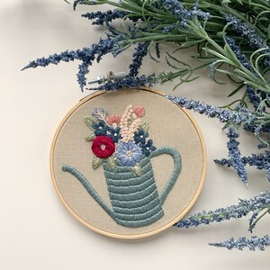 Op de afbeelding: Borduurkunst in een hoepel met een blauwe gieter gevuld met roze, blauwe en rode bloemen. De hoepel is van hout en heeft een beige stoffen achtergrond.