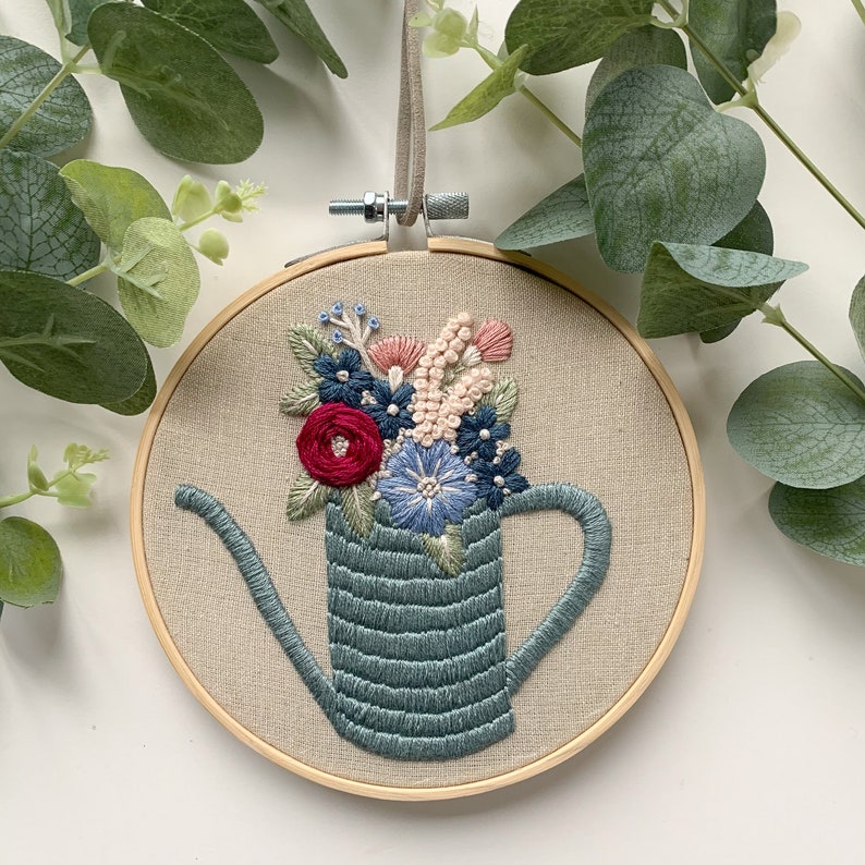 Op de afbeelding: Een handgestikte borduurring met een blauwe gieter gevuld met roze, blauwe en rode bloemen. De ring is gemaakt van natuurlijk hout en heeft een beige stoffen achtergrond.