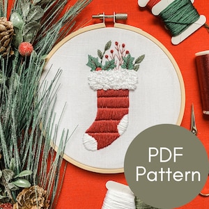May include: Embroidered Christmas stocking with red and white fabric, green holly leaves, and red berries. The stocking is in a wooden embroidery hoop on a red background. The text "PDF Pattern" is on a green circle.