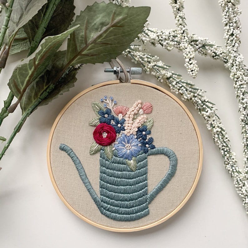 Op de afbeelding: Borduurwerk in een ronde hoepel met een blauwe gieter gevuld met een boeket roze, rode en blauwe bloemen.