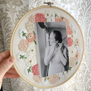 May include: A white embroidery hoop with a black and white photo of a person inside. The hoop is decorated with pink and white embroidered flowers.