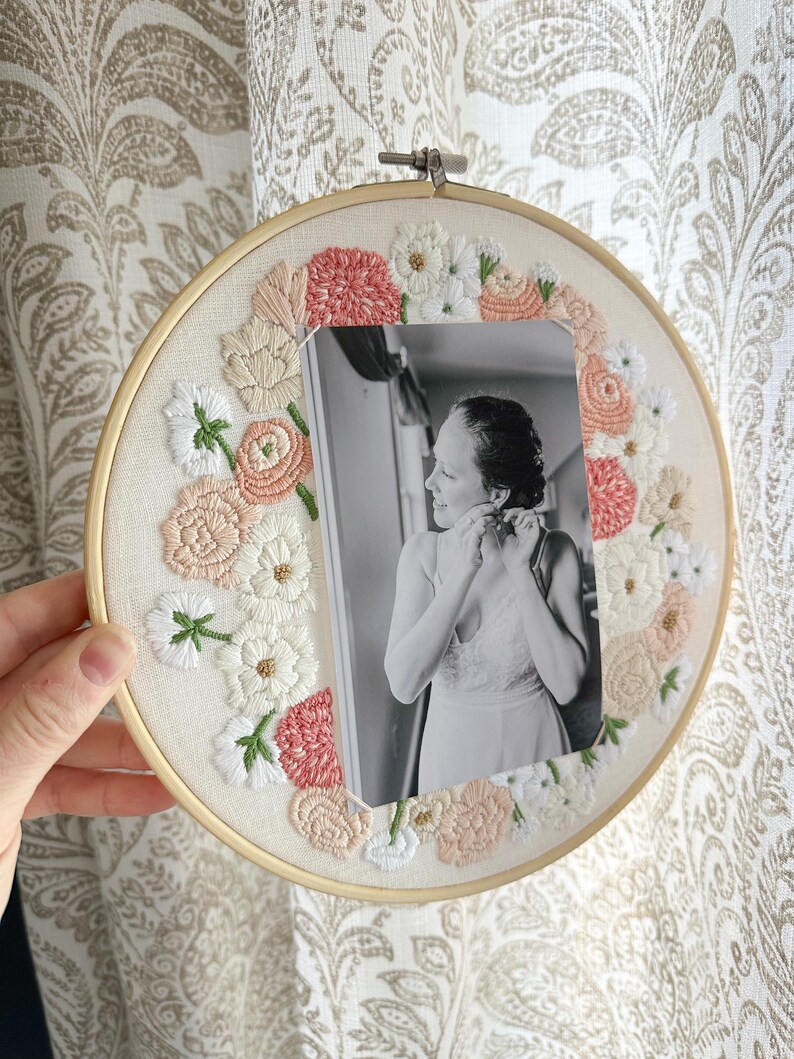 May include: A white embroidery hoop with a black and white photo of a person inside. The hoop is decorated with pink and white embroidered flowers.