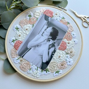 May include: A black and white photo of a person is framed with a hoop of embroidered fabric. The fabric is white with pink and white flowers.