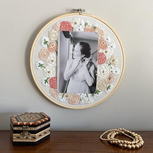 May include: A white embroidery hoop with a floral design in shades of pink and white. A black and white photo of a person is centered in the hoop.