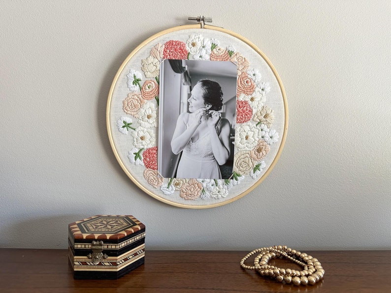 May include: A white embroidery hoop with a floral design in shades of pink and white. A black and white photo of a person is centered in the hoop.