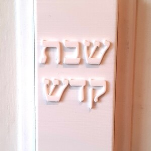 May include: A rectangular, light pink plaque with raised Hebrew lettering. The top line reads "Shabbat" and the bottom line reads "Kodesh". The plaque is mounted on a white wall.