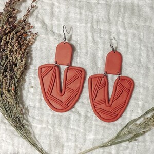 May include: A pair of terracotta-colored clay earrings with a geometric pattern. The earrings are shaped like a U with a smaller rectangle at the top. The earrings are hanging from silver hooks.