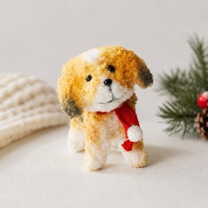 May include: A plush toy dog with a white and orange fur coat, black eyes, and a red scarf with a white pom-pom. The dog is standing on a white surface, with a knitted hat and pine needles in the background.