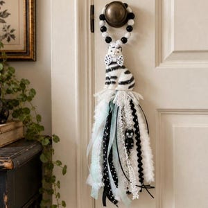 May include: A decorative door hanger featuring a ceramic black and white striped cat figurine with a polka dot bow tie. The cat sits atop a long, layered tassel made of various white, black, and mint green ribbons, lace, and a small cat charm.