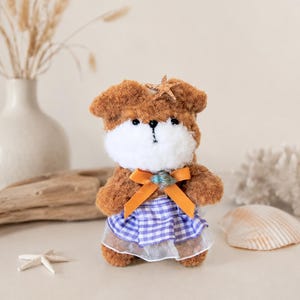 May include: A plush brown and white dog toy wearing a blue and white gingham dress with a white tulle underskirt. The dog has a starfish on its head and an orange bow around its neck. A small seashell is attached to the bow.